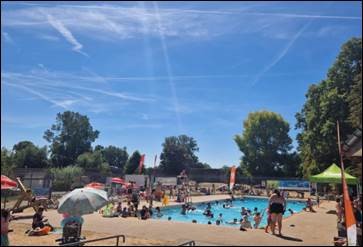 An image of Riverside Park pool in Wallingford summer 2022 with lots of people using the pool and walking around the facilities. The sky is blue with white wispy clouds