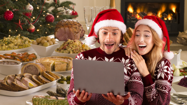 A pic of a couple dressed in xmas hats looking at a lap top with Xmas food in the background